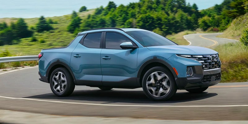 Blue Hyundai Santa Cruz pickup truck driving on a scenic road surrounded by green hills.