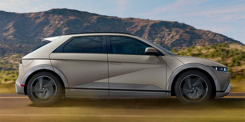 Silver Hyundai IONIQ 5 electric vehicle driving on a scenic road with mountains in the background.