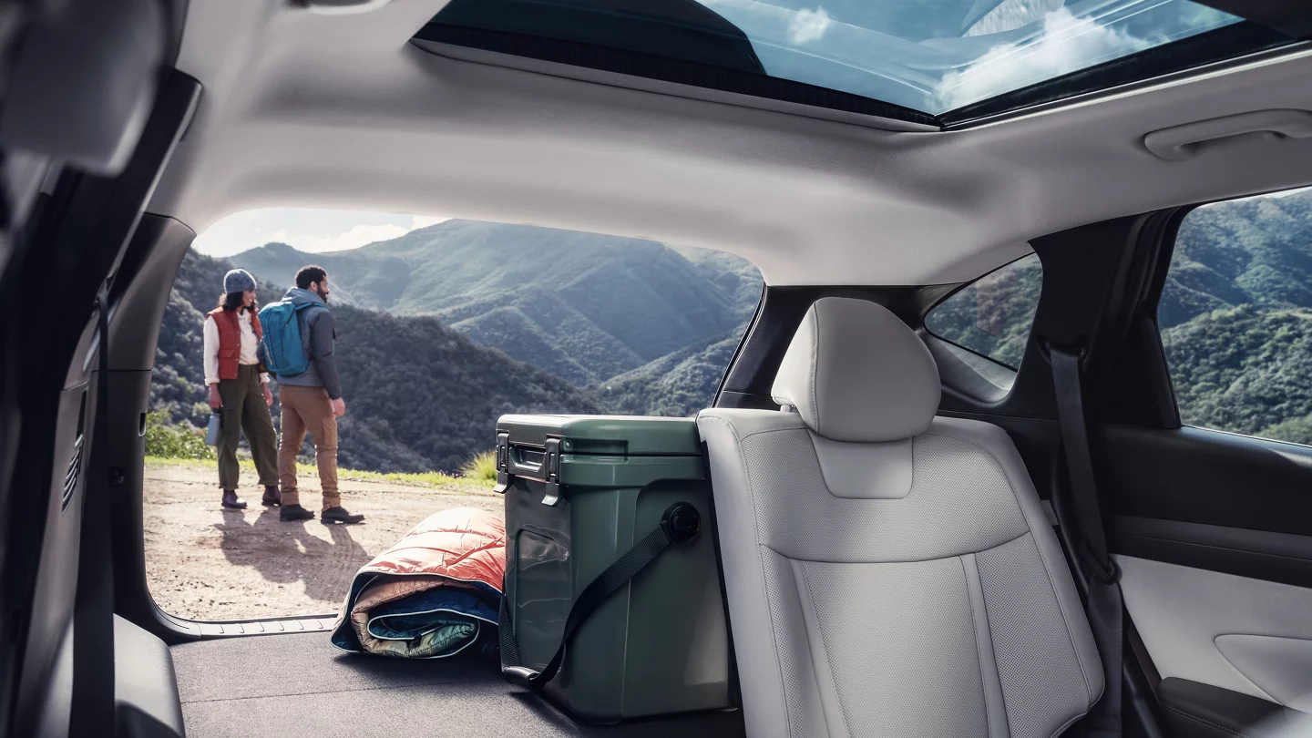 View from the back of a 2025 Hyundai Tucson with a gray interior, featuring a panoramic sunroof and a green cooler with a rolled-up sleeping bag in the trunk. A couple in hiking gear stands outside, looking at scenic mountain views. Available at Windward Hyundai.
