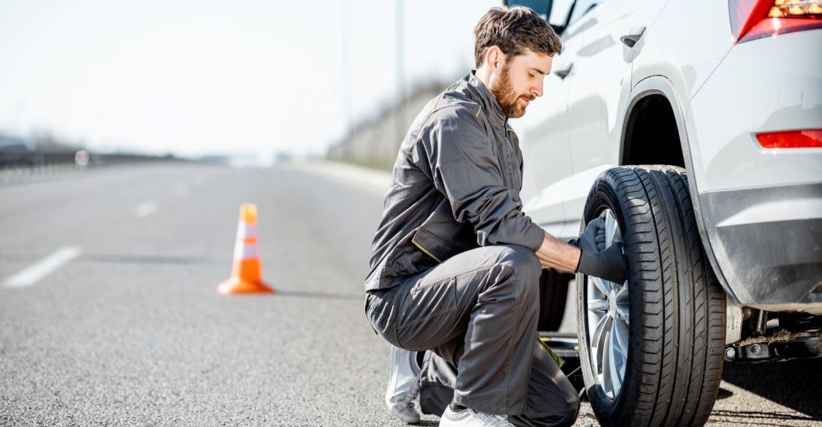 Roadside technician changing a flat tire on a white SUV, with safety cones placed for protection on a highway shoulder.