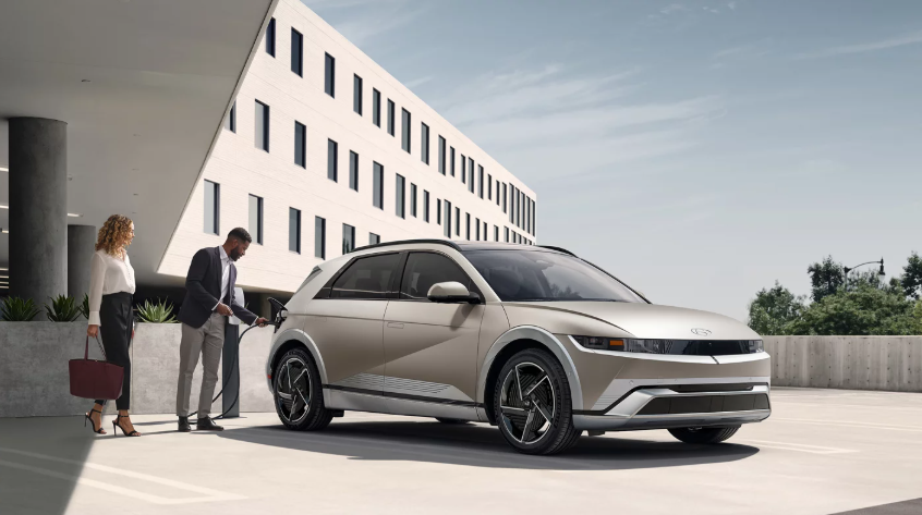 Silver 2026 Hyundai IONIQ 5 electric SUV charging outside modern office building with two people standing nearby.